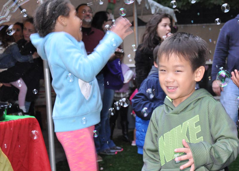 black girl dancing in bubbles, Asian boy in hoodie in the foreground smiling